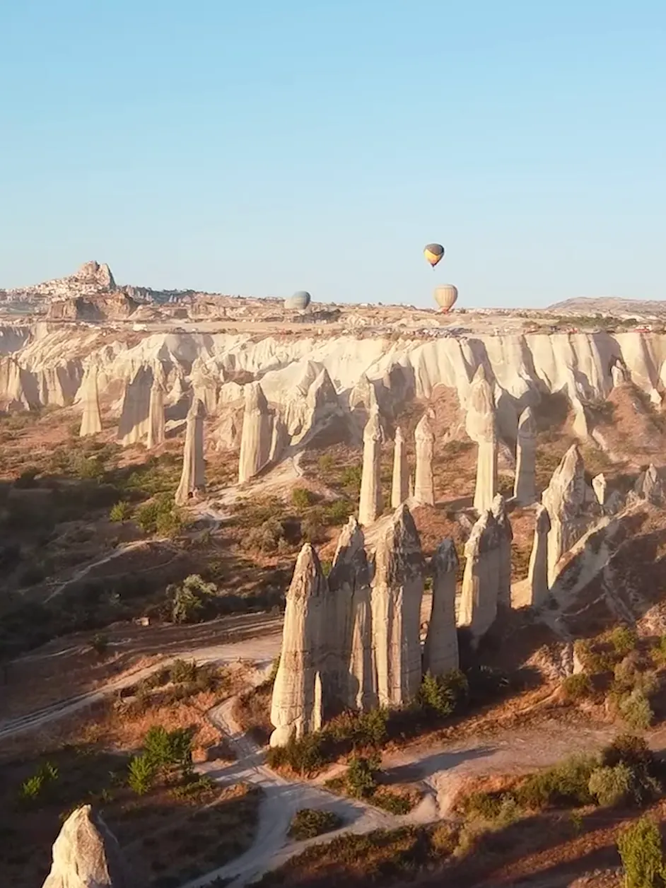 Rock pinnacles of a Cappadocian valley at first light, Uçhisar on the horizon and three balloons far off