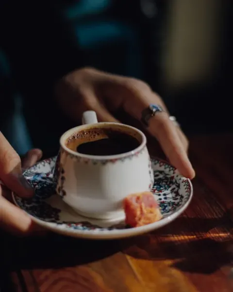 Turkish coffee served in a small cup, held in one hand, with a glass of water and a piece of lokum